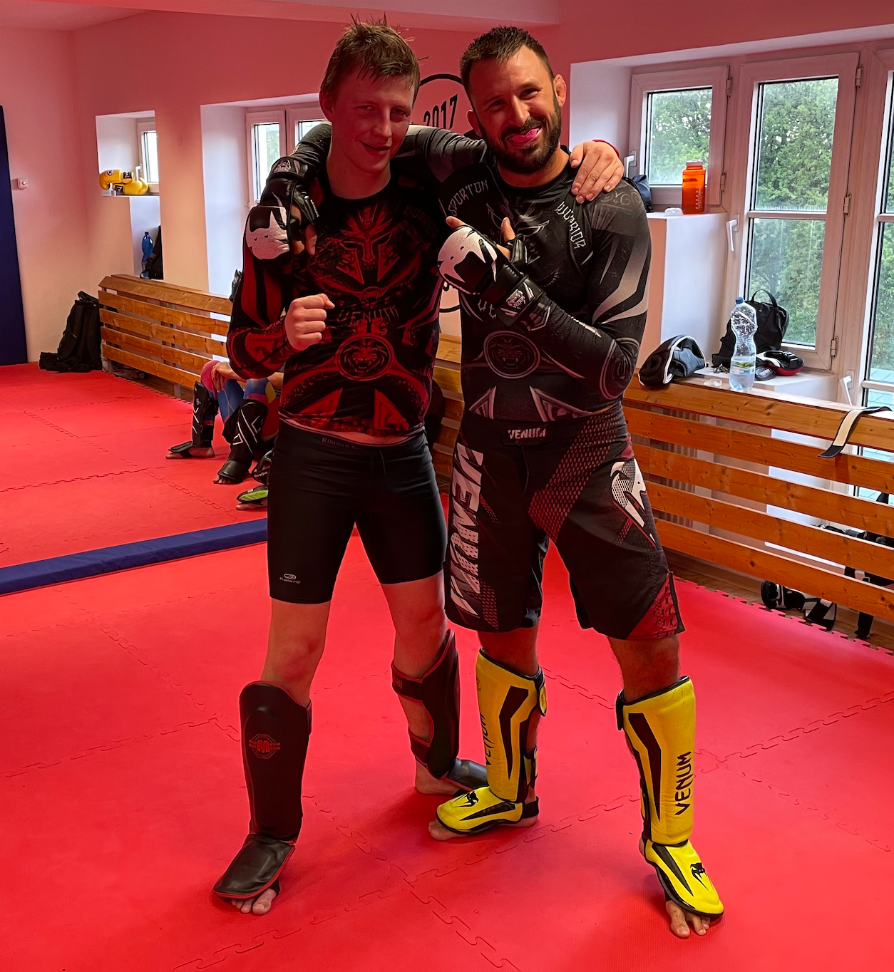 Two training partners wearing combat-sports gloves and shin guards in a gym.